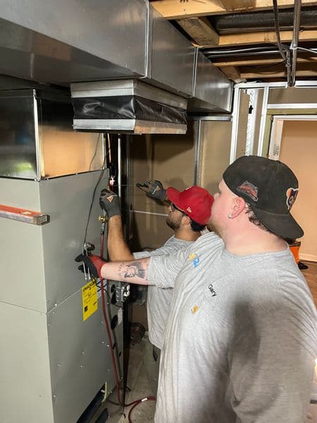 Technicians servicing HVAC system in basement with tools and equipment.