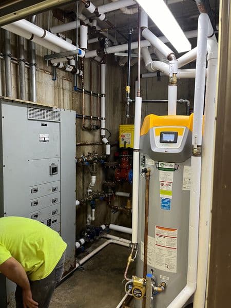 Worker servicing a water heater in a utility room with exposed plumbing and electrical panel.