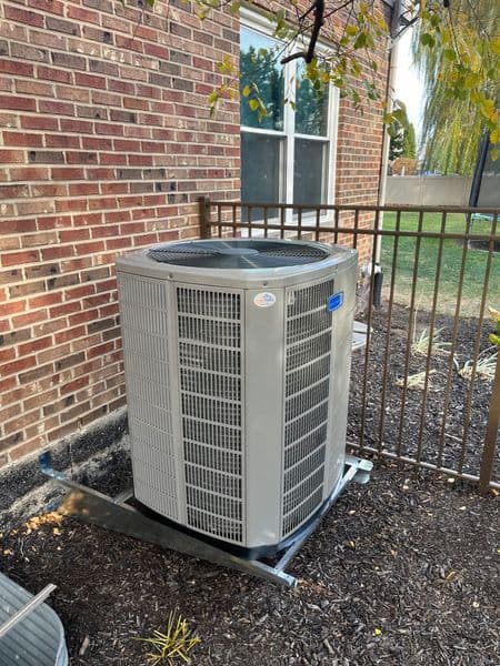 Modern air conditioning unit installed outside a brick house, surrounded by mulch and a fence.