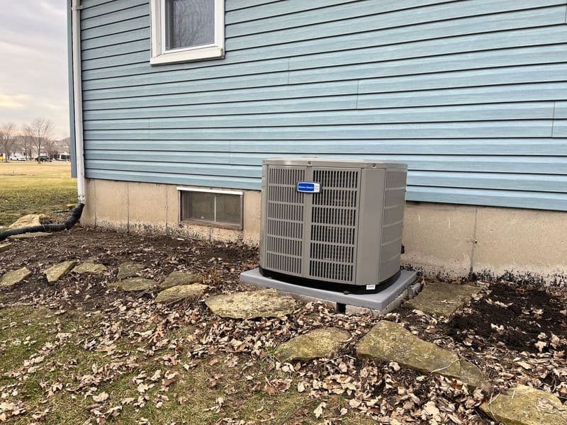Air conditioning unit installed outside a light blue house, surrounded by stones and leaves.