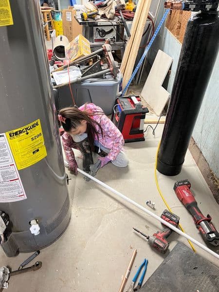 Young girl working on plumbing in a cluttered garage with tools and water heater visible.