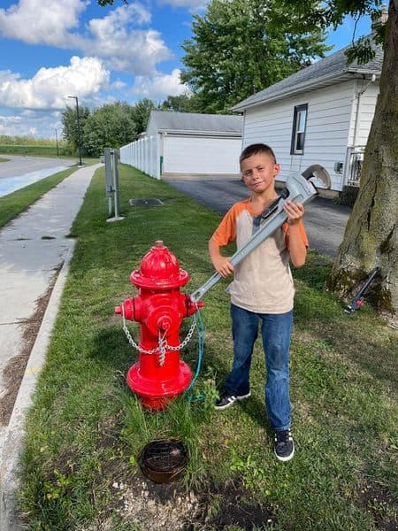 Young boy holding a large wrench next to a bright red fire hydrant on a sunny day.