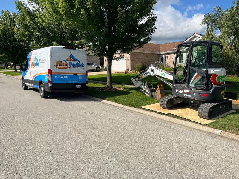 Excavator and service van parked on a residential street, surrounded by trees and homes.