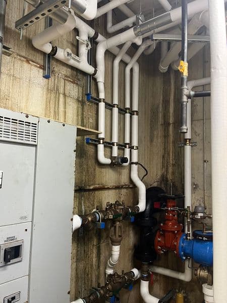 Complex plumbing system with white PVC pipes and valves against a textured wall.