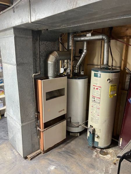 Furnace and water heater in a basement with exposed pipes and concrete walls.