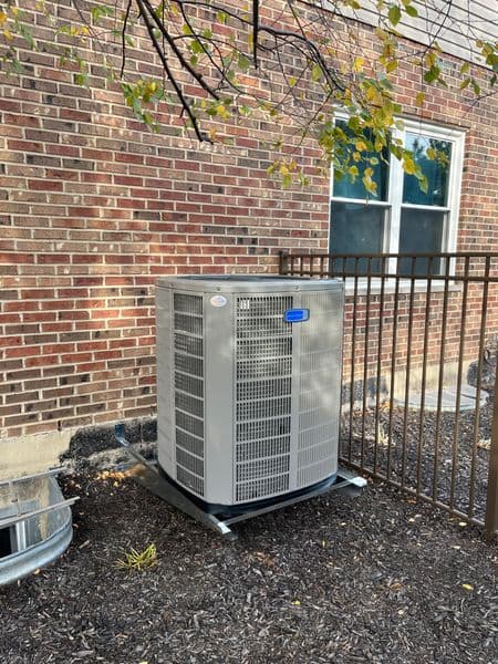 Air conditioning unit installed beside brick building, surrounded by landscaping and fencing.
