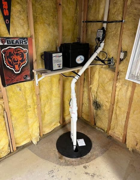 Home sump pump installation with pipe, battery backup, and support shelf in a shed.